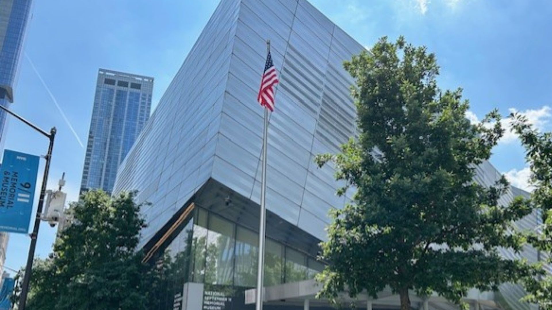 Exterior of the 9/11 Memorial & Museum against a blue sky, with green trees and an American flag on a flagpole at the foreground