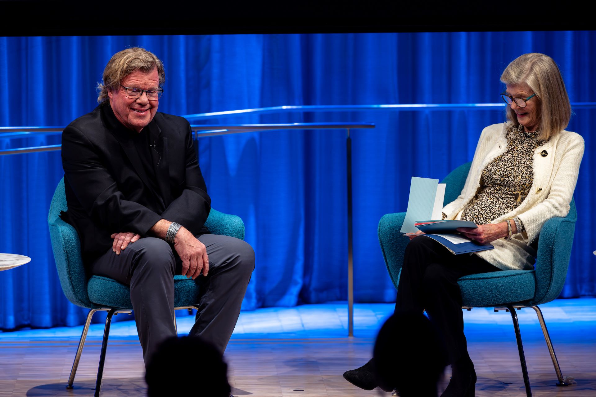 Joe McNally (left) and Jan Ramirez (right) sit on stage in front of a blue curtain, smiling and talking.