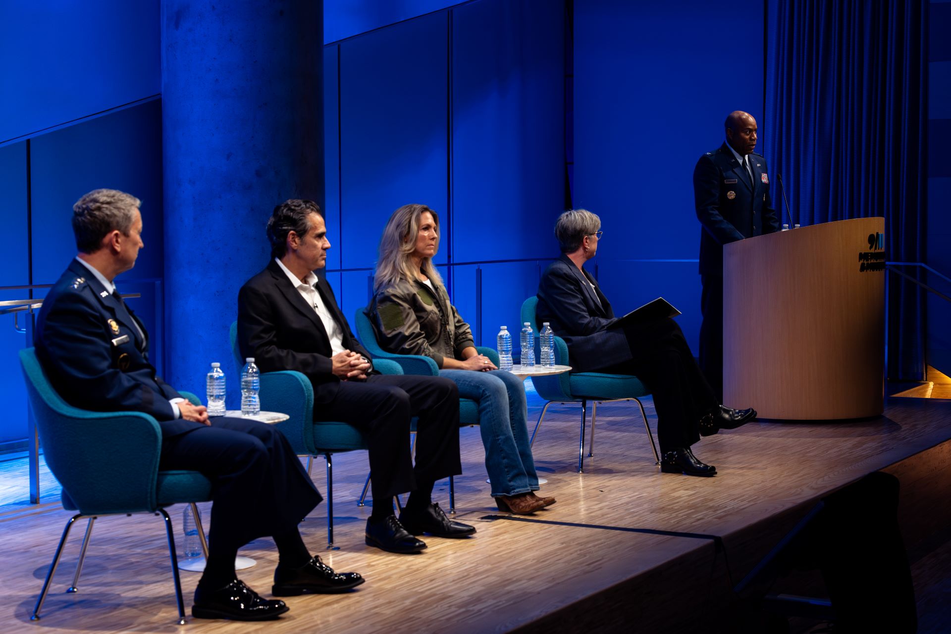 a row of four seated panelists face a podium on stage where a person introduces the event