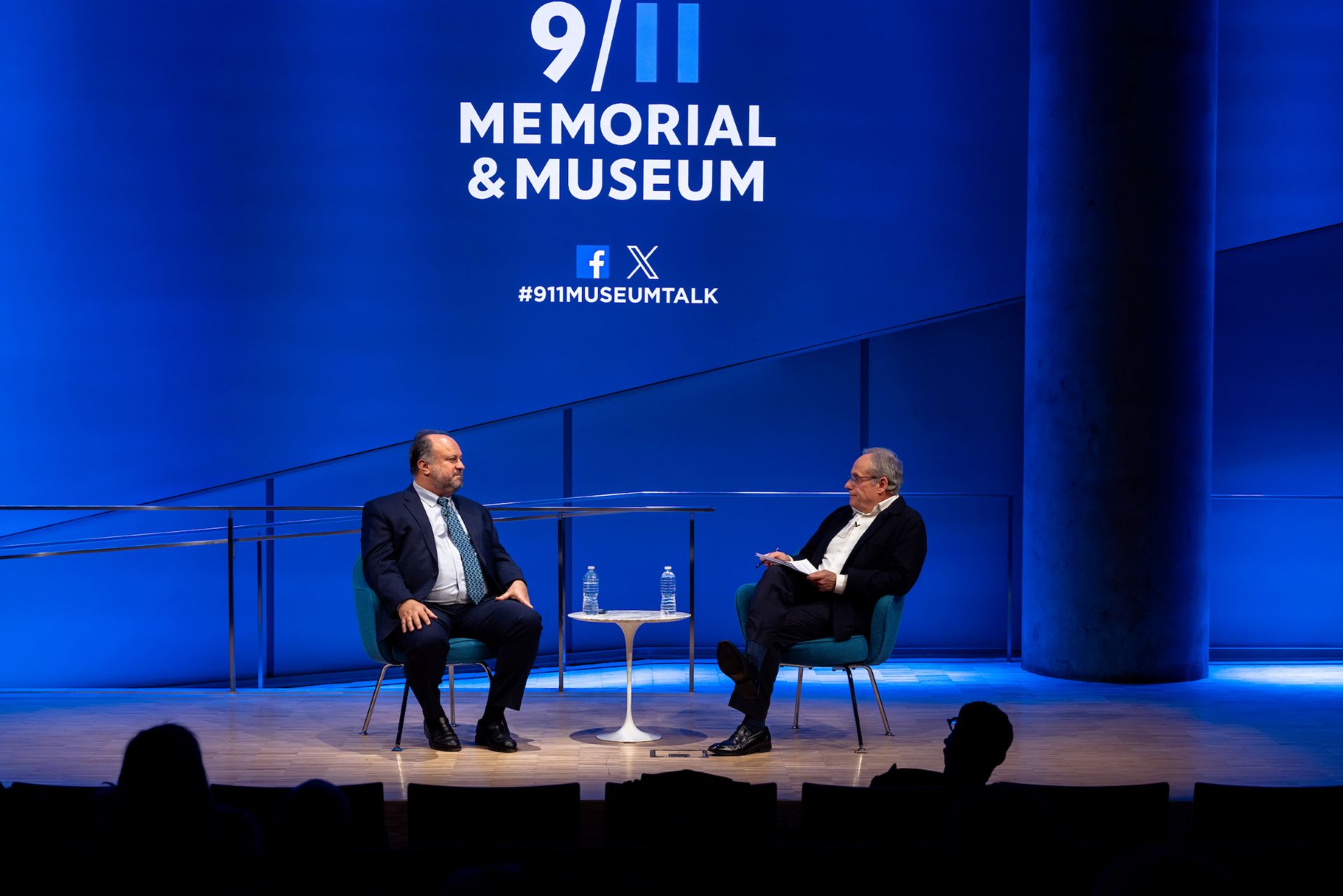 Bernard Haykel and Cliff Channing sit on a sage in two chairs, facing each other across a table. 9/11 Memorial & Museum's logo is visible on a blue wall behind them