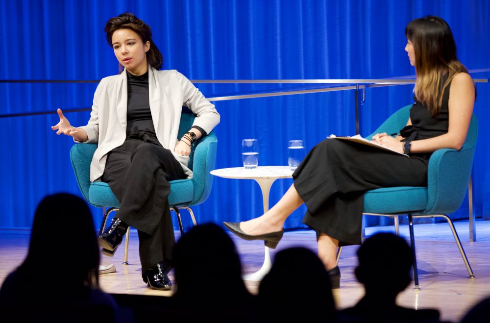 VICE correspondent Isobel Yeung looks at the audience as she speaks onstage at the Museum Auditorium. She is gesturing with her left hand and her legs are crossed. A woman hosting the event listens as she sits with her legs crossed.