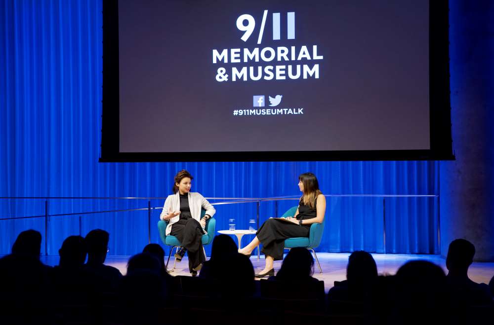 VICE correspondent Isobel Yeung speaks onstage in this wide-angle photo from the audience. A woman hosting the event sits beside her and listens as she speaks. Audience members are silhouetted by lights onstage. A projector with the 9/11 Memorial & Museum logo has been lowered above them.
