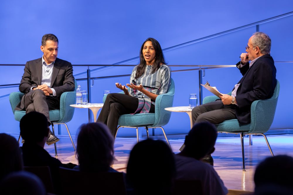 Vidhya Ramalingam, founder of Moonshot CVE, holds out her hands as she speaks onstage at the Museum Auditorium. Hany Farid, Dartmouth computer science professor and senior advisor to the Counter Extremism Project, listens with his hands in his lap to her left. Clifford Chanin, the executive vice president and deputy director for museum programs, sits to her right holding a clipboard and listening on. Audience members in the foreground are silhouetted by the white and blue lights onstage.