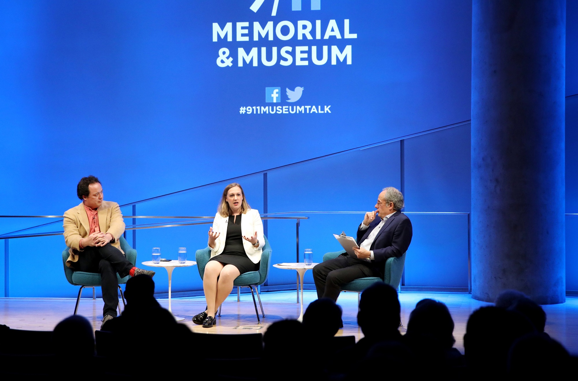 A wide-angle photo of the Museum Auditorium stage shows Devorah Margolin of George Washington University, Graeme Wood of The Atlantic and and Clifford Chanin, the executive vice president and deputy director for museum programs, taking part in a public program. Margolin is seated at the center of the stage gesturing with both hands as she speaks to the audience. Wood is to her right with his hands on his knee and Chanin is to her left holding a clipboard. Audience members are silhouetted in the foreground.