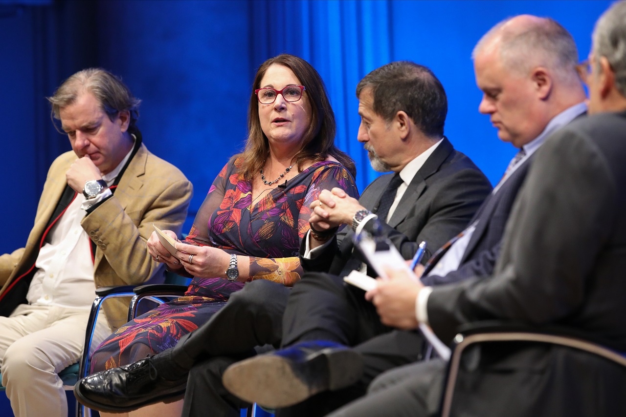 This close-up photo shows four men and one woman speaking onstage at the Museum Auditorium during a public program. Al-Qaeda expert Mary E. Galligan speaks while looking out at the audience, who are not visible. She is holding a piece of paper in her hands. Al-Qaeda experts Peter Bergen and Mark Stout are to her left and right, respectively. Museum advisor Mark Stout and Clifford Chanin, the executive vice president and deputy director for museum programs, are seated to her left as well. Chanin is somewhat 