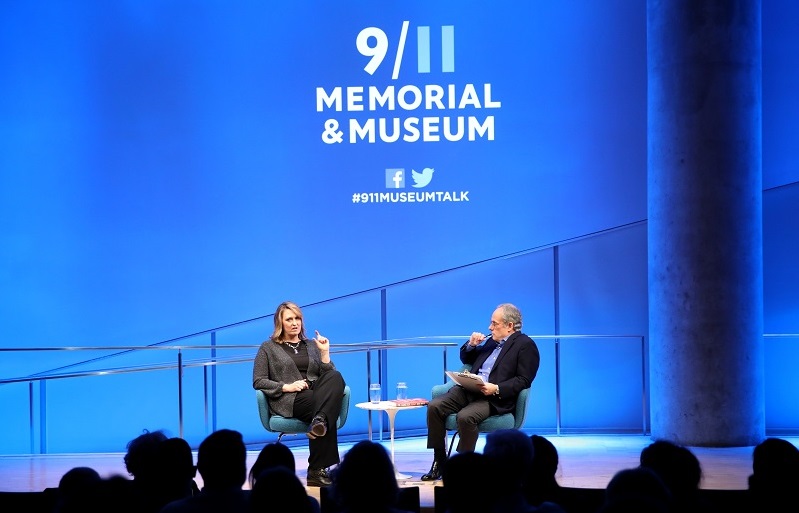 In this photo from behind the audience, Author and former CIA analyst and targeting officer Nada Bakos and Clifford Chanin, the executive vice president and deputy director for museum programs, take part in a public program in the Museum Auditorium. The two of them are seated onstage as a silhouetted audience watches on. The bright white and blue lights of the stage fall on the wall behind them. The logo of the 9/11 Memorial & Museum is projected on the wall.