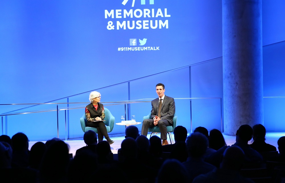 In this wide-angle of the Museum Auditorium’s stage, Boston Globe reporter Mitchell Zuckoff and a woman hosting a public program sit next to each other in front of an audience. Zuckoff is speaking while looking out at the audience as the woman looks on with her legs crossed and her hands in her lap. The 9/11 Memorial & Museum logo is projected above them. Blue and white lights shine on and behind the stage. The silhouettes of dozens of audience members are in the foreground.