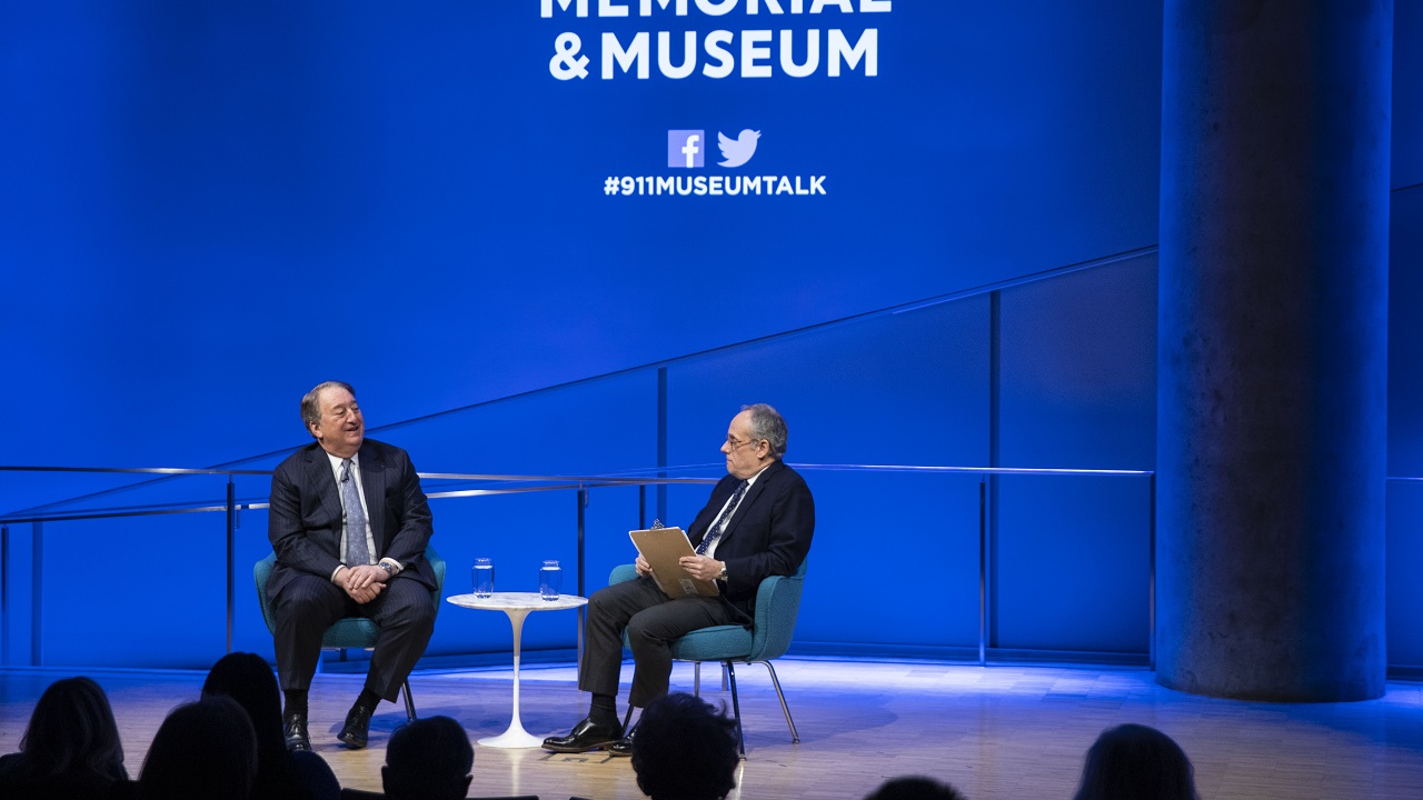 In this wide-angle view from the audience, real estate owner and builder Howard Milstein and moderator Clifford Chanin sit onstage during a public program at the Museum Auditorium. Milstein is speaking with his hands in his lap as Chanin listens while holding a clipboard. The blue lights of the stage shine on the wall behind them. The heads of audience members are silhouetted in the foreground.