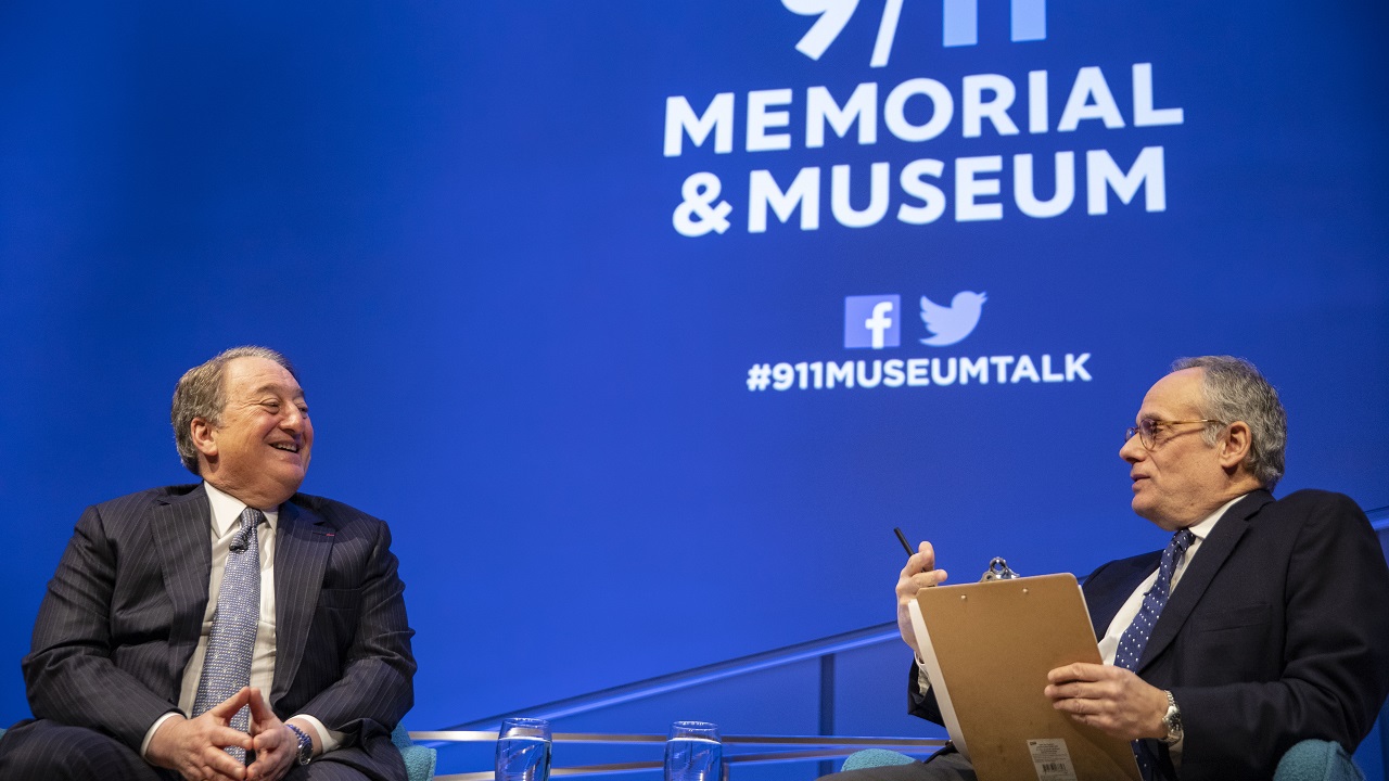 In this close-up view of real estate owner and builder Howard Milstein and moderator Clifford Chanin, the two men look at each other as they’re seated onstage at the Museum Auditorium. Chanin is holding a clipboard in one hand and a pen in his other hand. Milstein is smiling with his hands in his lap. The 9/11 Memorial & Museum logo is projected on a wall behind them.