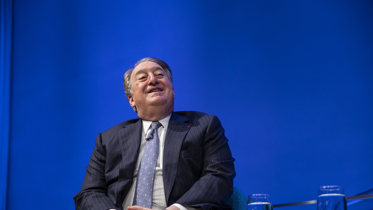 Real estate owner and builder Howard Milstein smiles as he sits in a chair on the Auditorium stage in this close-up view. The wall behind him is lit up blue.