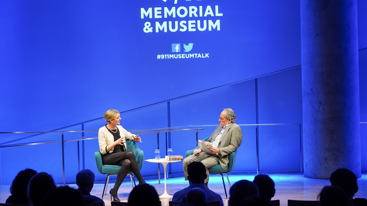 In this view from the audience, author Joana Cook gestures with both hands as she speaks with moderator Clifford Chanin. The silhouetted heads of audience members are in the foreground as the two interact onstage. Chanin is holding a clipboard as he looks at Cook. The 9/11 Memorial & Museum logo is projected on a blue-lit wall behind them.