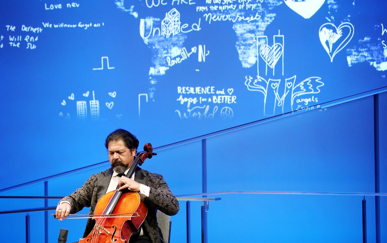 Cellist Karim Wasfi performs with eyes closed while seated onstage in the Museum Auditorium. His bright orange-brown cello contrasts with the blue lights of the stage.
