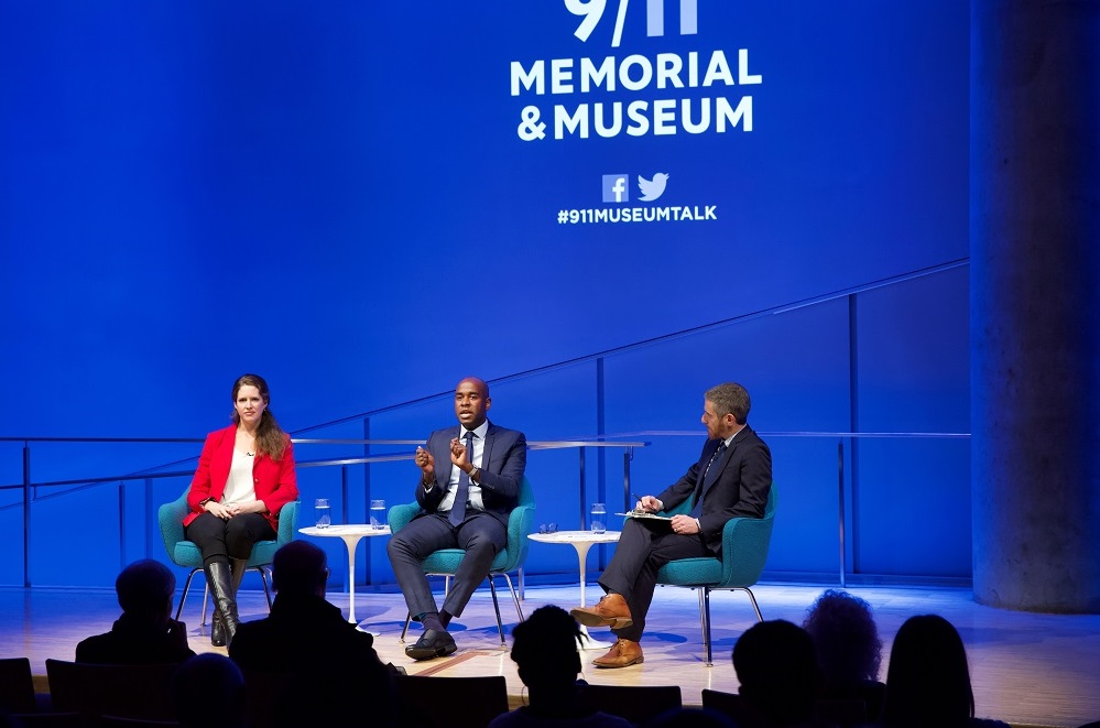 Three public program participants sit on a blue-lit auditorium stage in this wide-angle photograph. Above their heads, “9/11 Memorial & Museum #museumtalk” is projected onto the wall behind them. On the far left, a woman in a red blazer looks off into the audience. In the center, a man gestures with his hands and out into the audience. The moderator sits with a clipboard in his lap and looks at the man speaking. The heads of the audience members appear in the foreground in silhouette.