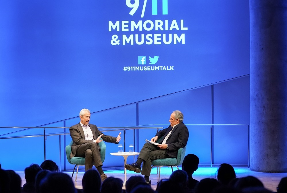 Two men in suits sit cross-legged on a blue-lit auditorium stage. "9/11 Memorial & Museum #museumtalk" is projected onto the wall behind them. The heads of the audience members appear in silhouette in front of them.