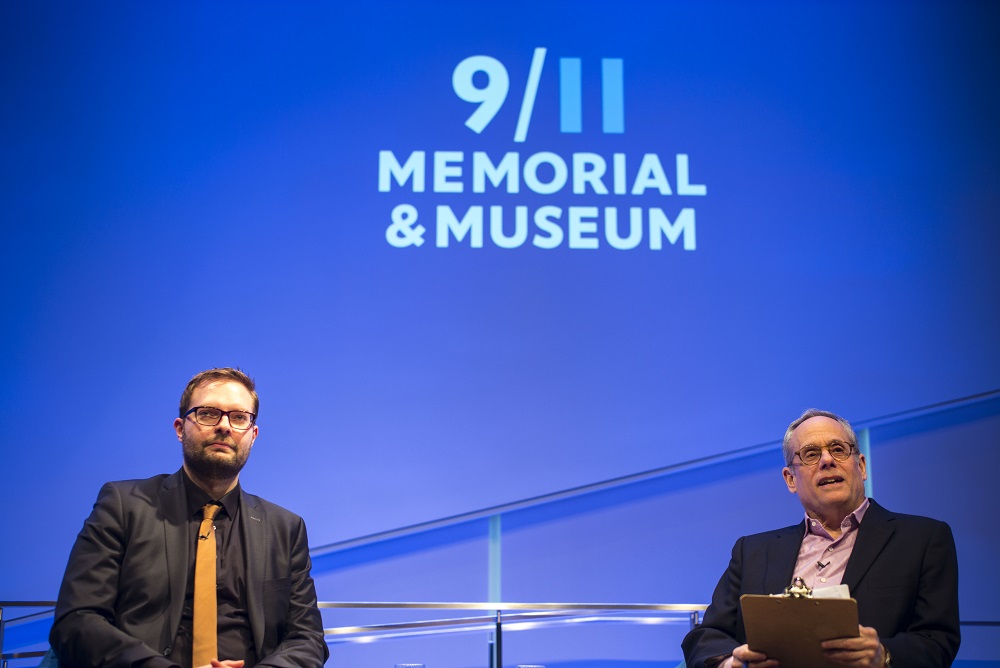 Two men in glasses, a public program participant and a moderator, sit on a blue-lit auditorium stage with "9/11 Memorial & Museum" projected onto the wall behind them. They both look out into an unseen audience.