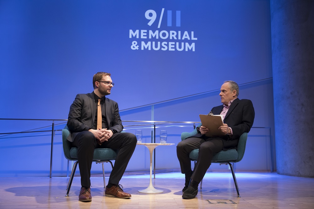 Two men in glasses, a public program participant and a moderator, sit on a blue-lit auditorium stage with "9/11 Memorial & Museum" projected onto the wall behind them. They address each other.