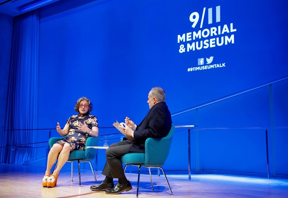 A public program participant sits on a blue-lit auditorium stage with a program moderator. "9/11 Memorial & Museum #museumtalk" is projected onto the wall behind them.