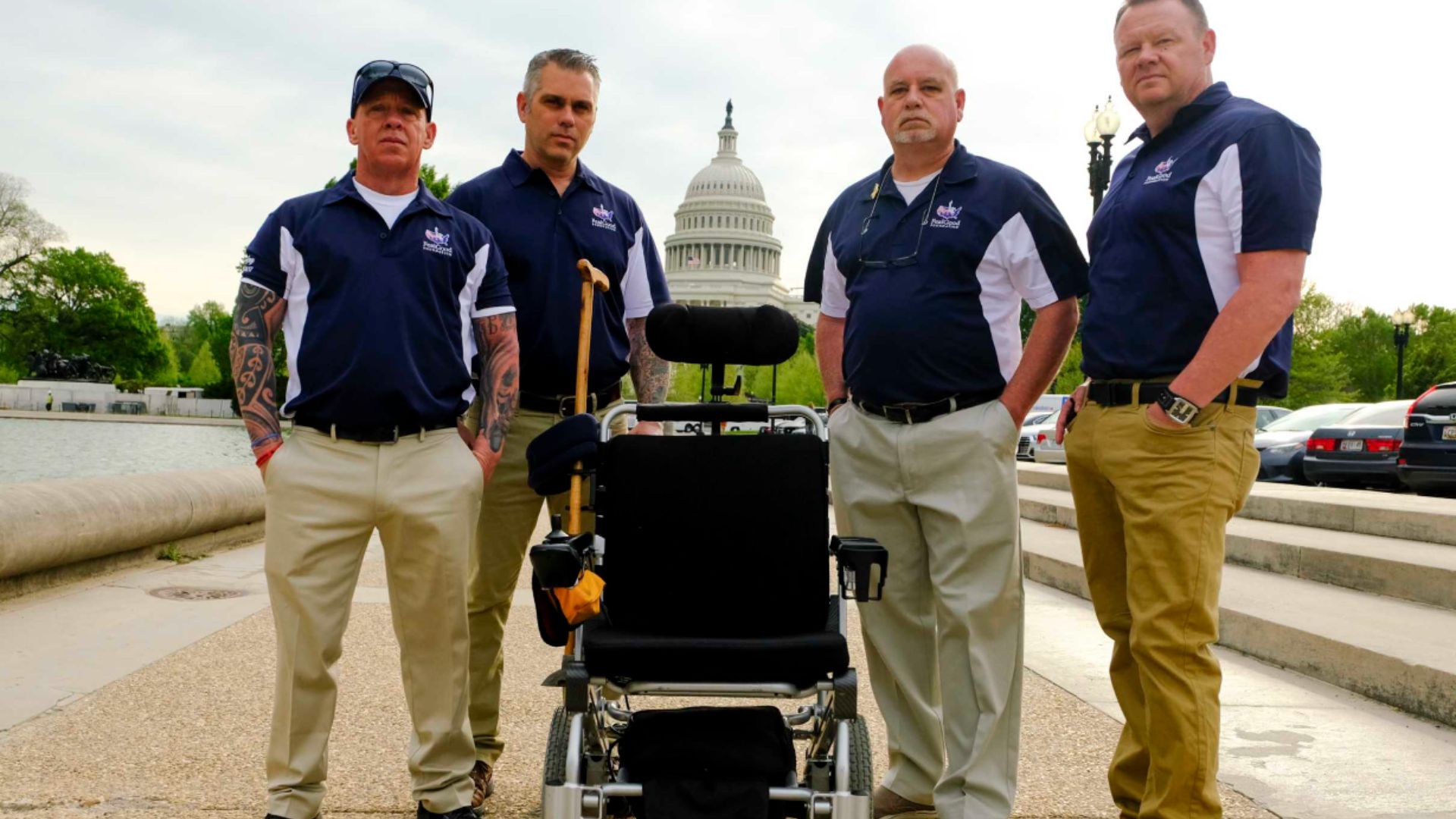 Four men in khaki pants and navy blue shirts stand with an empty wheelchair in front of the U.S. capital building.