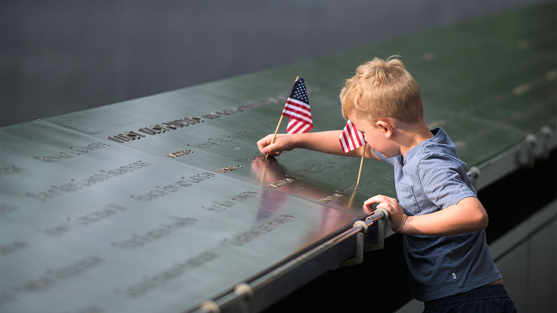 A small, light-haired child touches the 9/11 Memorial and a small American flag placed at one of the victim's names