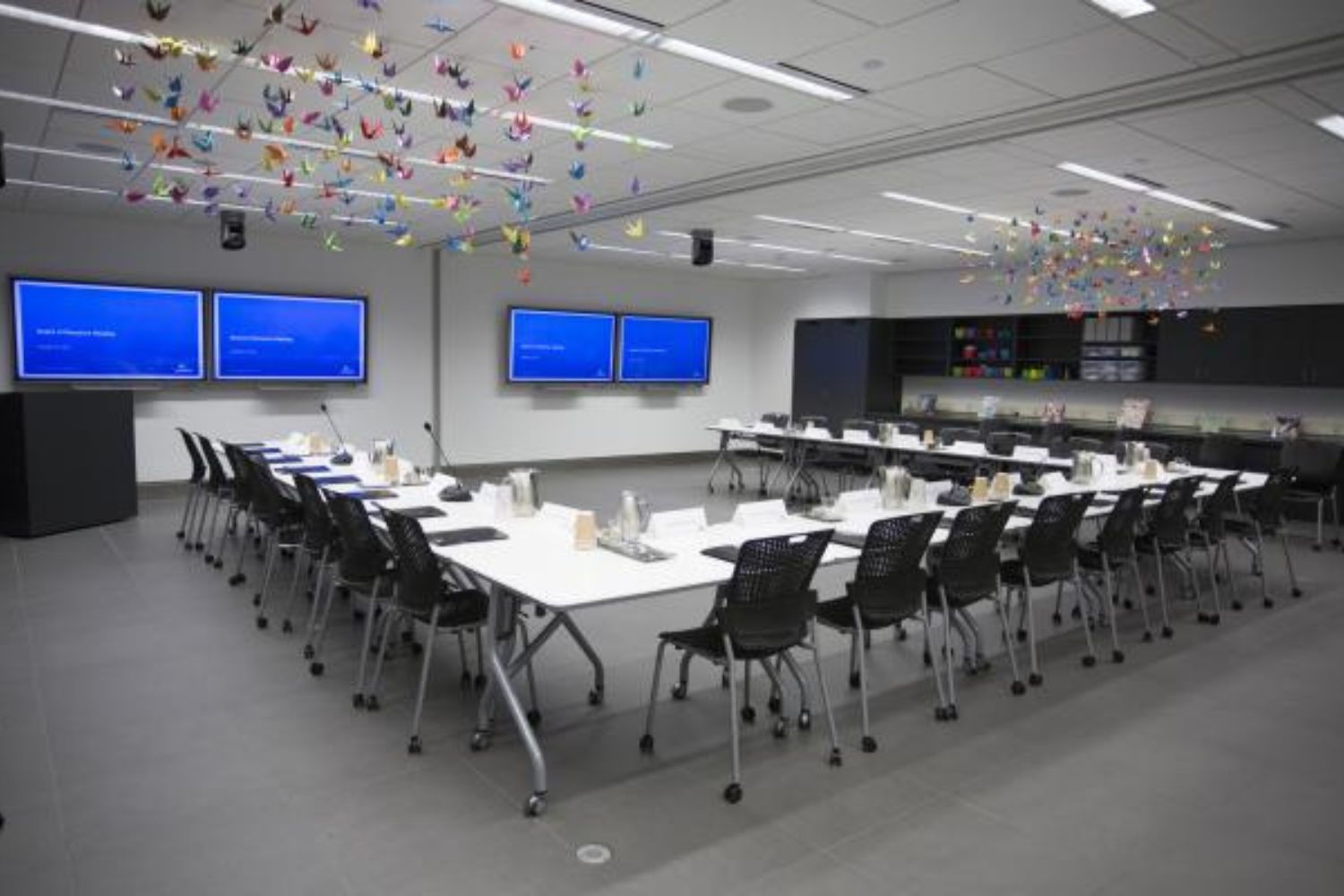 An open classroom, with bright white tables and lights facing blue screens. Oragami birds hang from ceiling