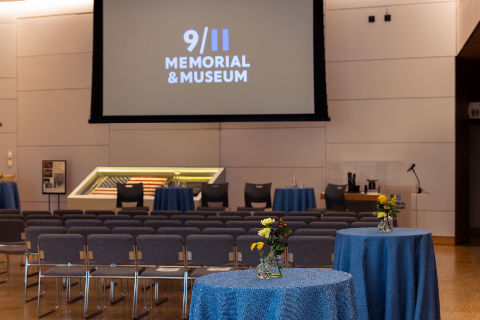 a screen shows the Museum logo with a stage set, chairs in the middle ground, and blue tables in the back