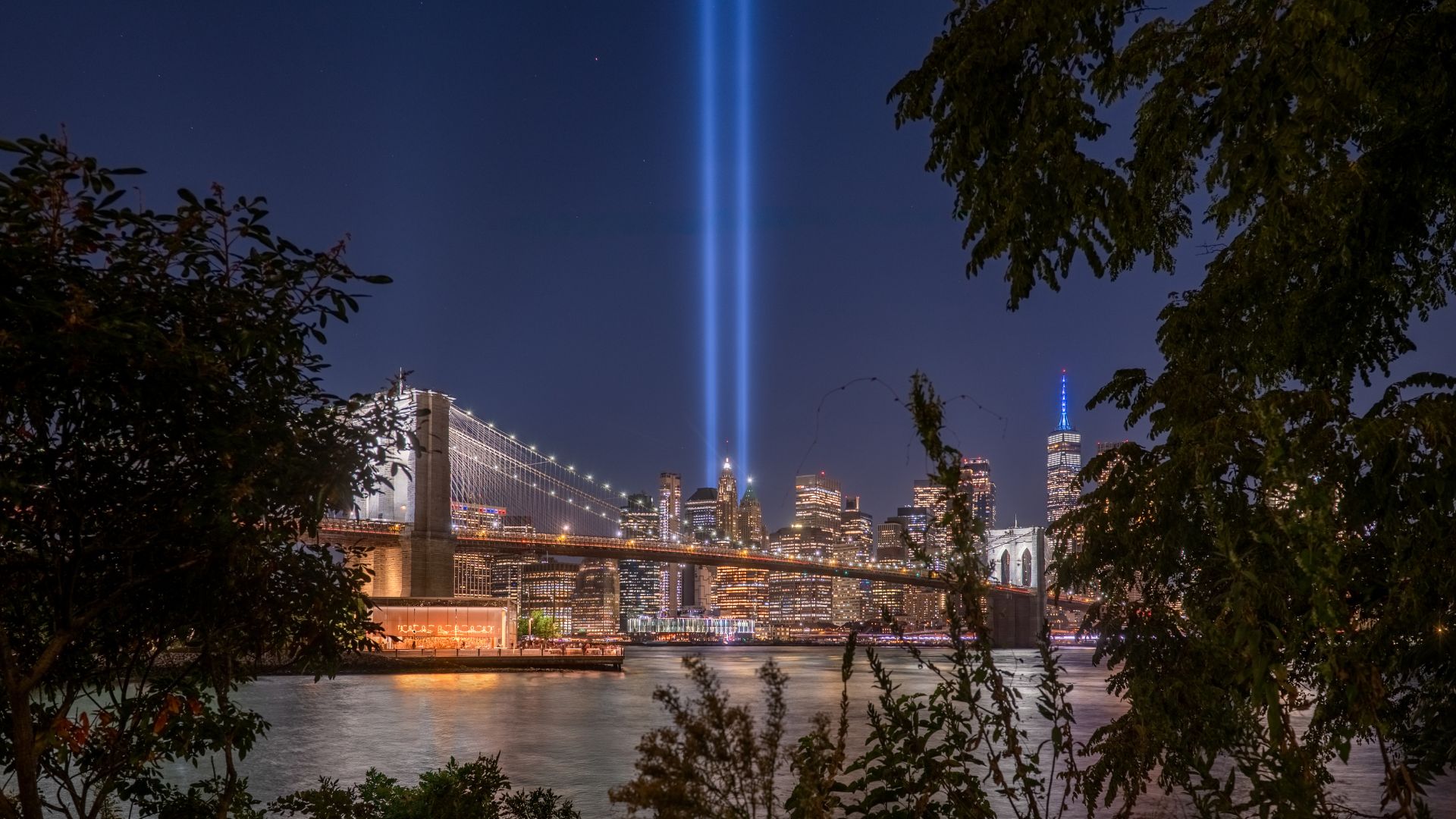 Twin beams of blue beams of light radiate into the nighttime sky with the New York City skyline in the background. 