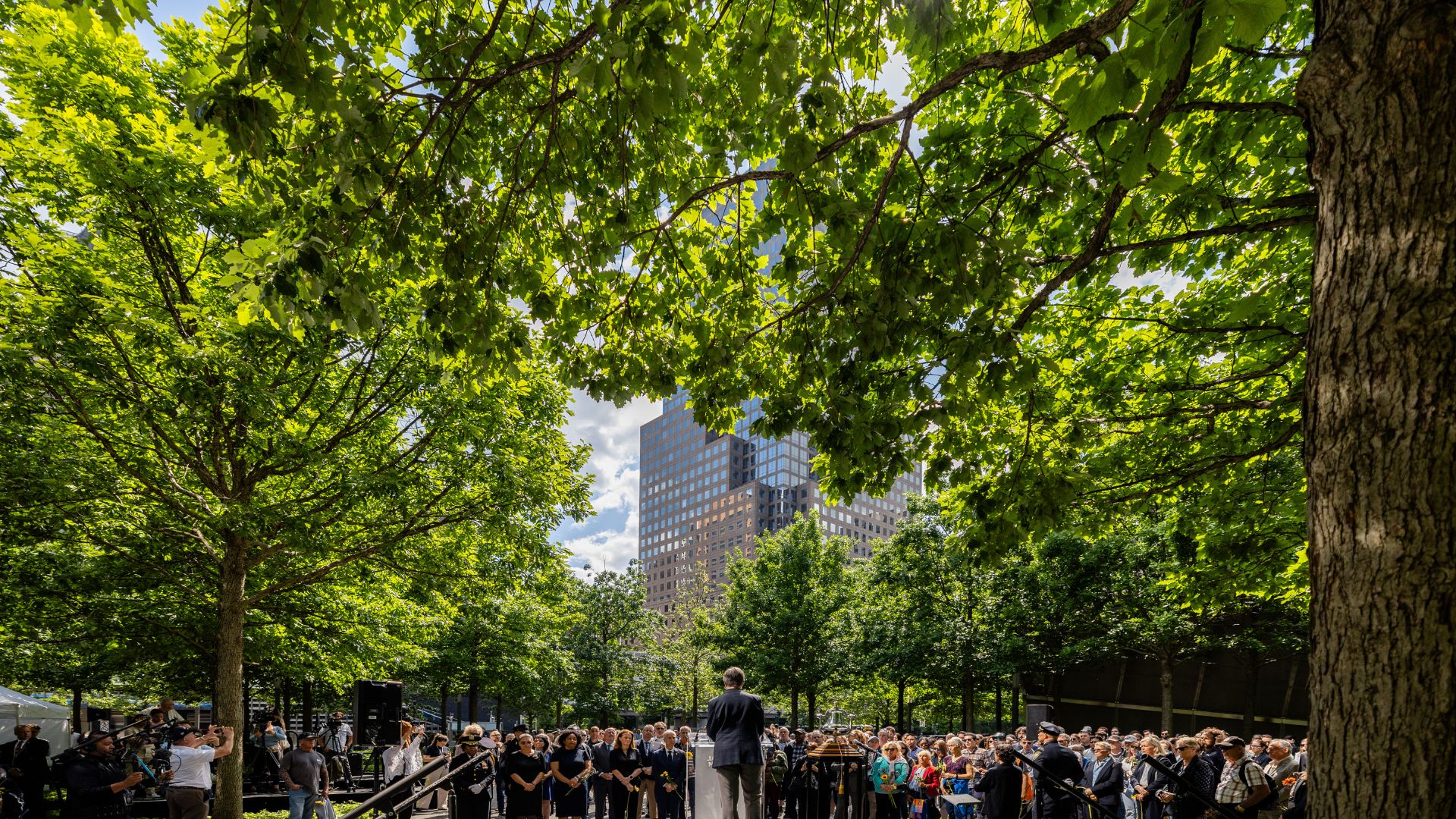 A crowd listens to a speaker against a backdrop of full green foliage on the Memorial plaza.