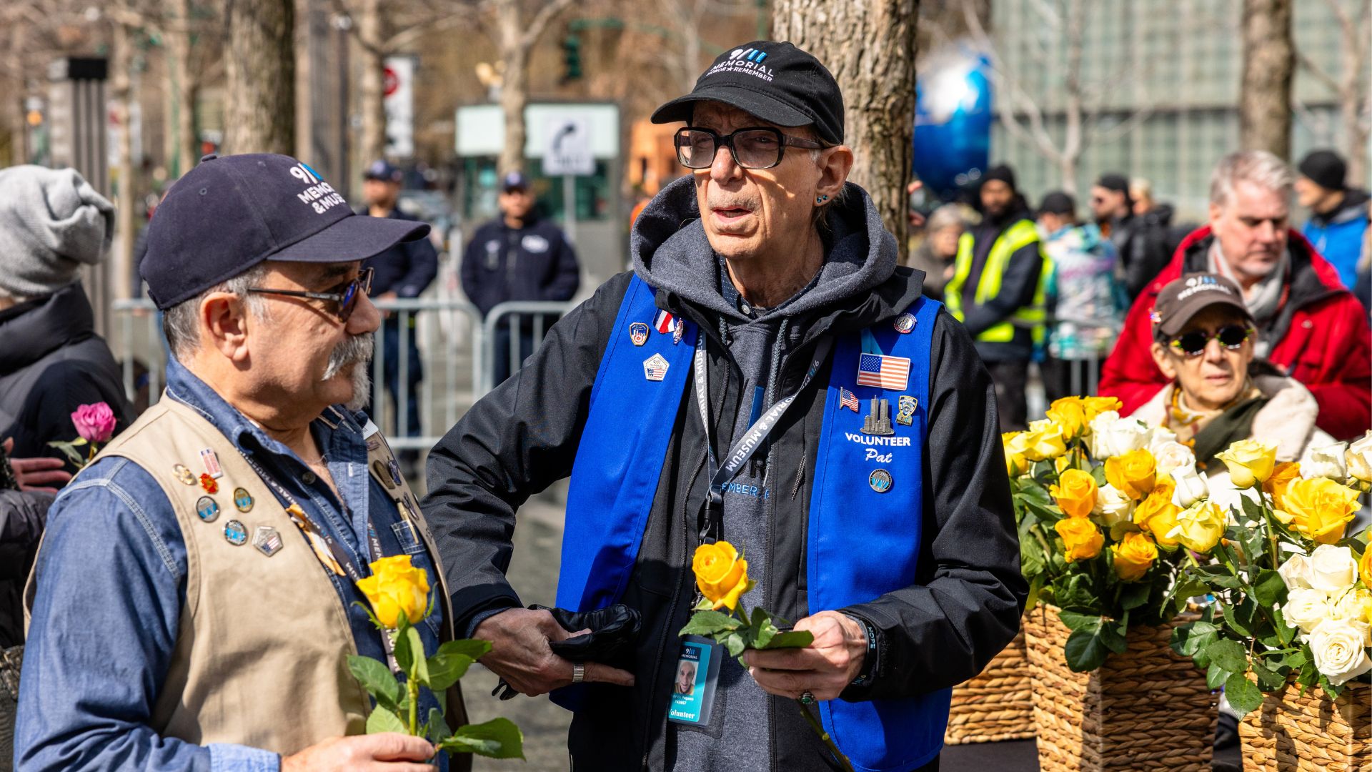 Two peole - one in a blue vest and one in a beige vest - speak to one another while holding yellow roses, with more in the background on the Memorial parapets.