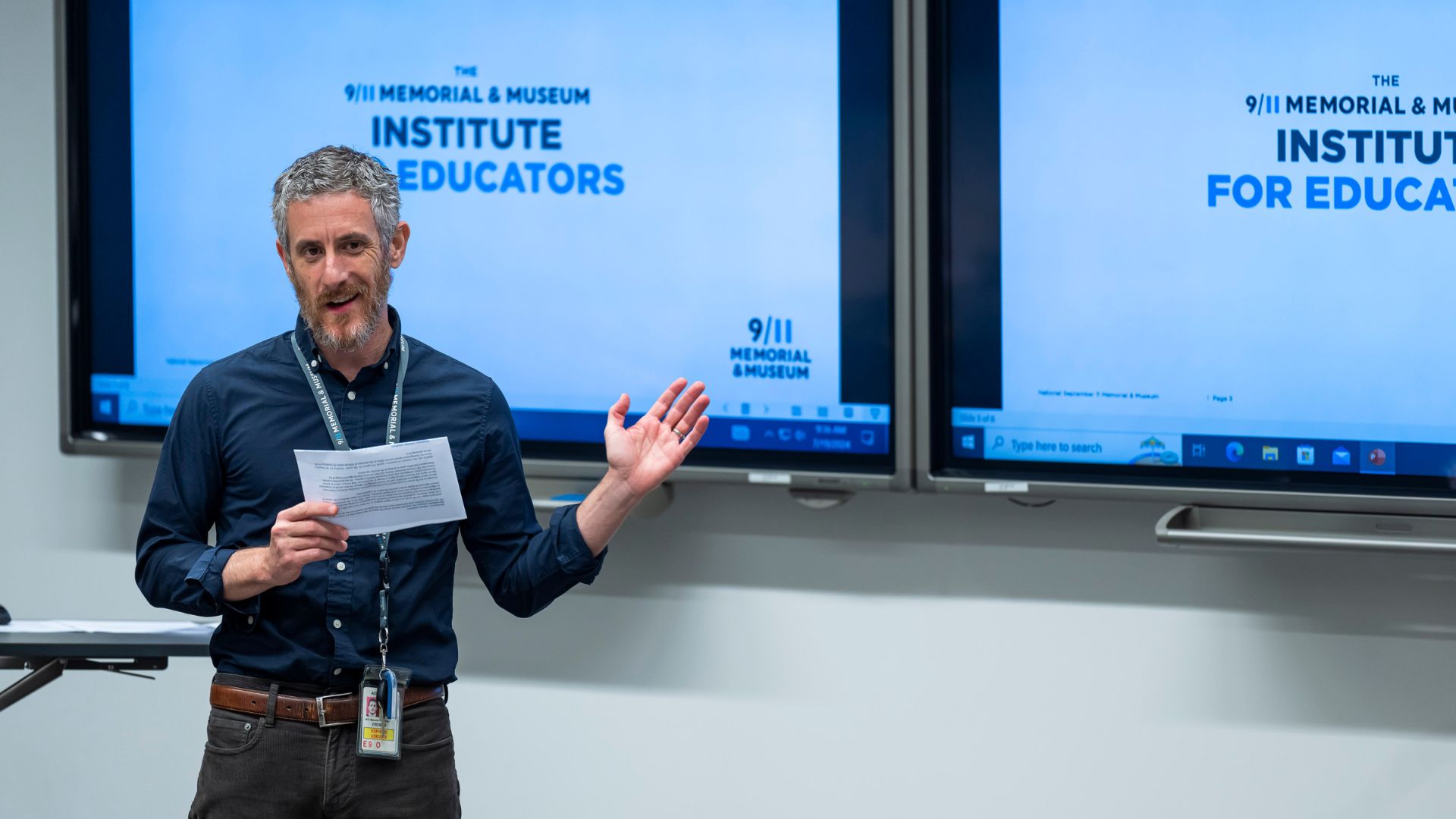 A person in a blue shirt and brown pants stands in front of two screens that read "9/11 Memorial & Museum Institute for Educators," while reading off a piece of paper and with one hand in the air.