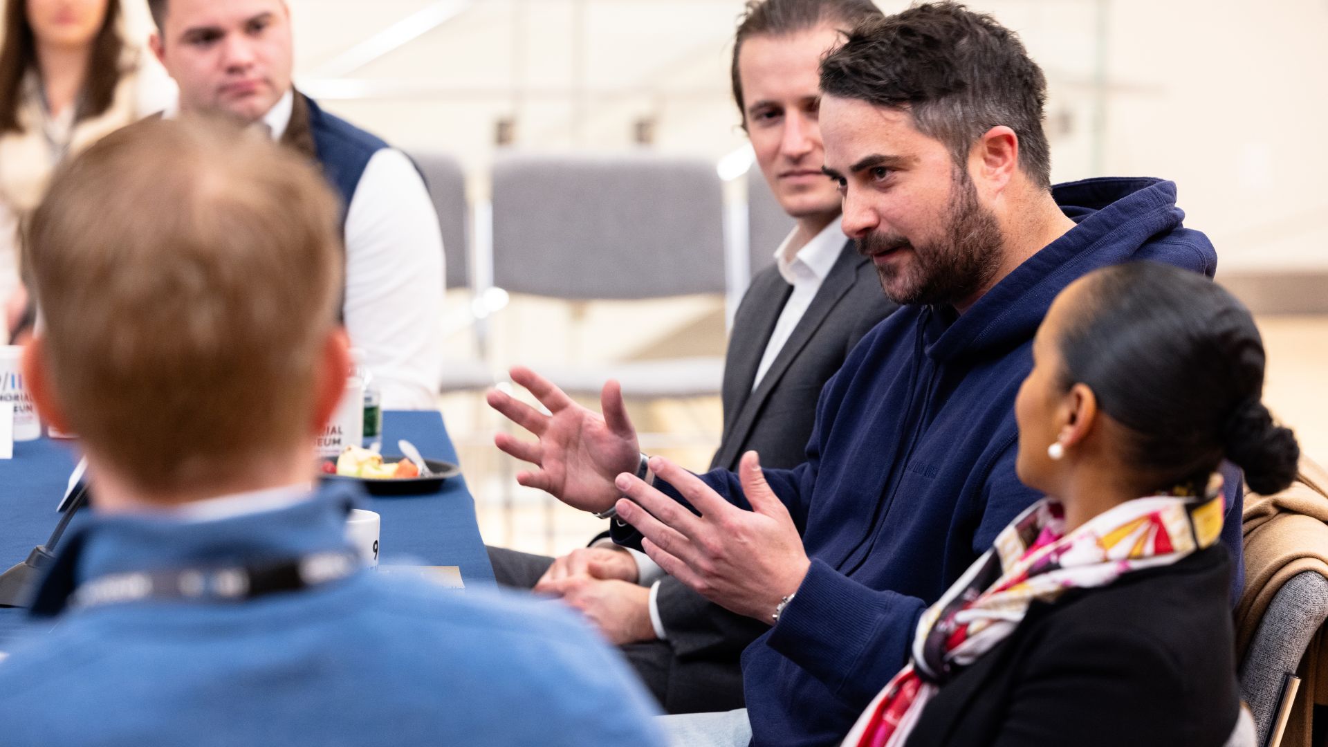 Five people seated at a table mid-discussion. A dark-haired person with a beard wearing a navy blue hoodie speaks animatedly with his hands.