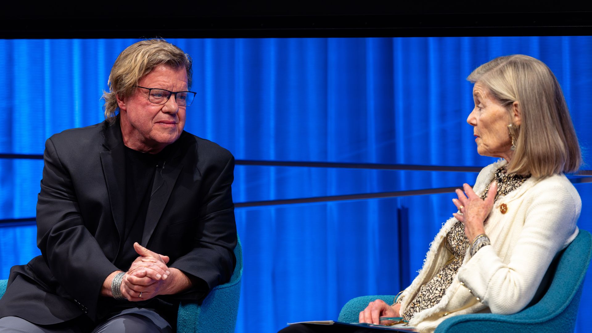 Two people in conversation on stage, against a bright blue curtain. The one on the left wears a dark suit. The one on the right wears a cream-colored sweater.