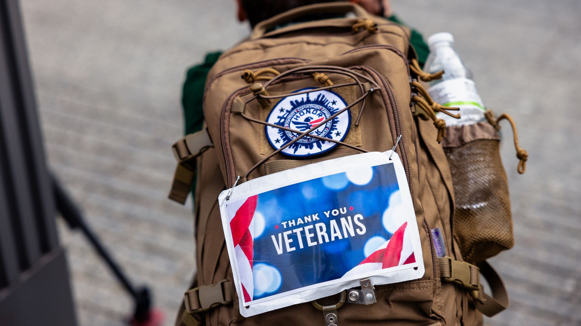 Back view of an individual with a tan-colored backpack bearing a round patch that says, "Honor Ruck" and a rectangular patch that says "Thank You Veterans."