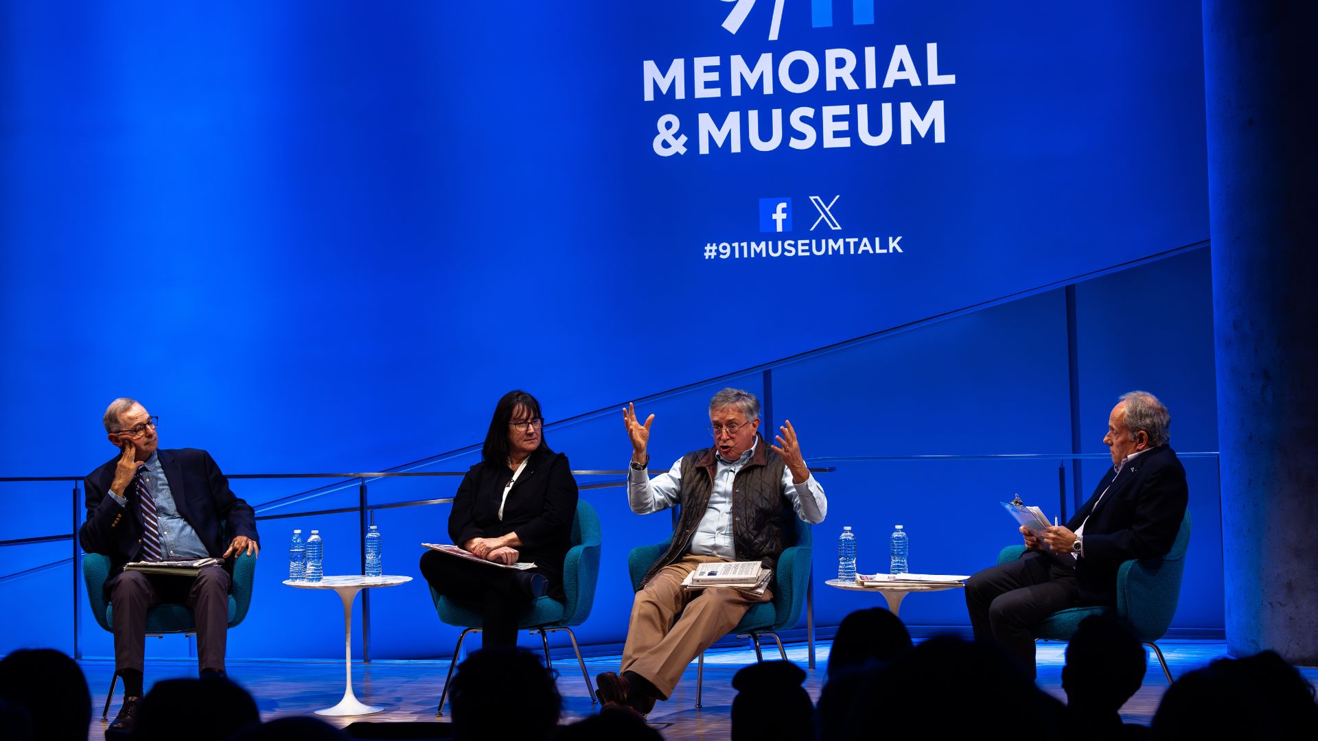 Four seated panelists in a discussion on stage, against a bright blue curtain adorned with the 9/11 Memorial & Museum logo.