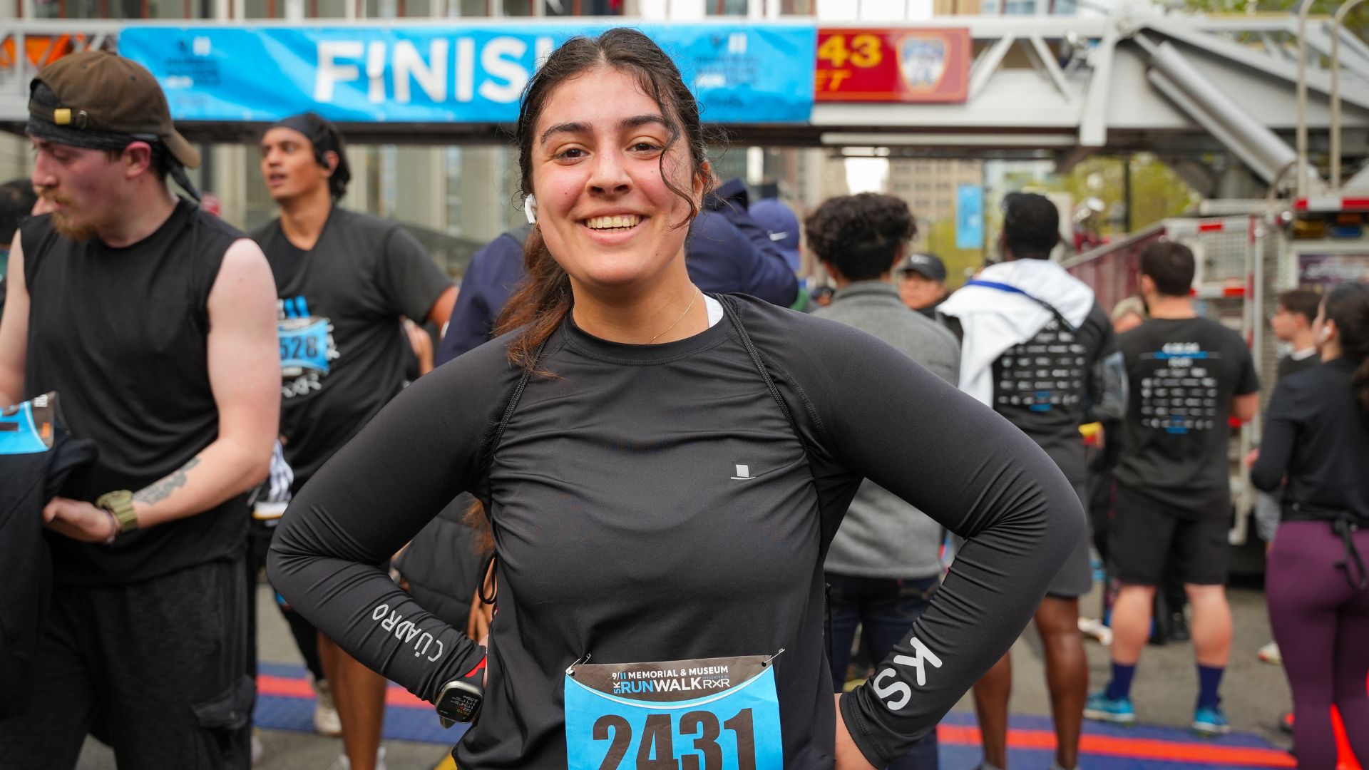 A runner stands with their hands on their hips at the finish line of the 5K. A crowd of other participants is visible in the background.