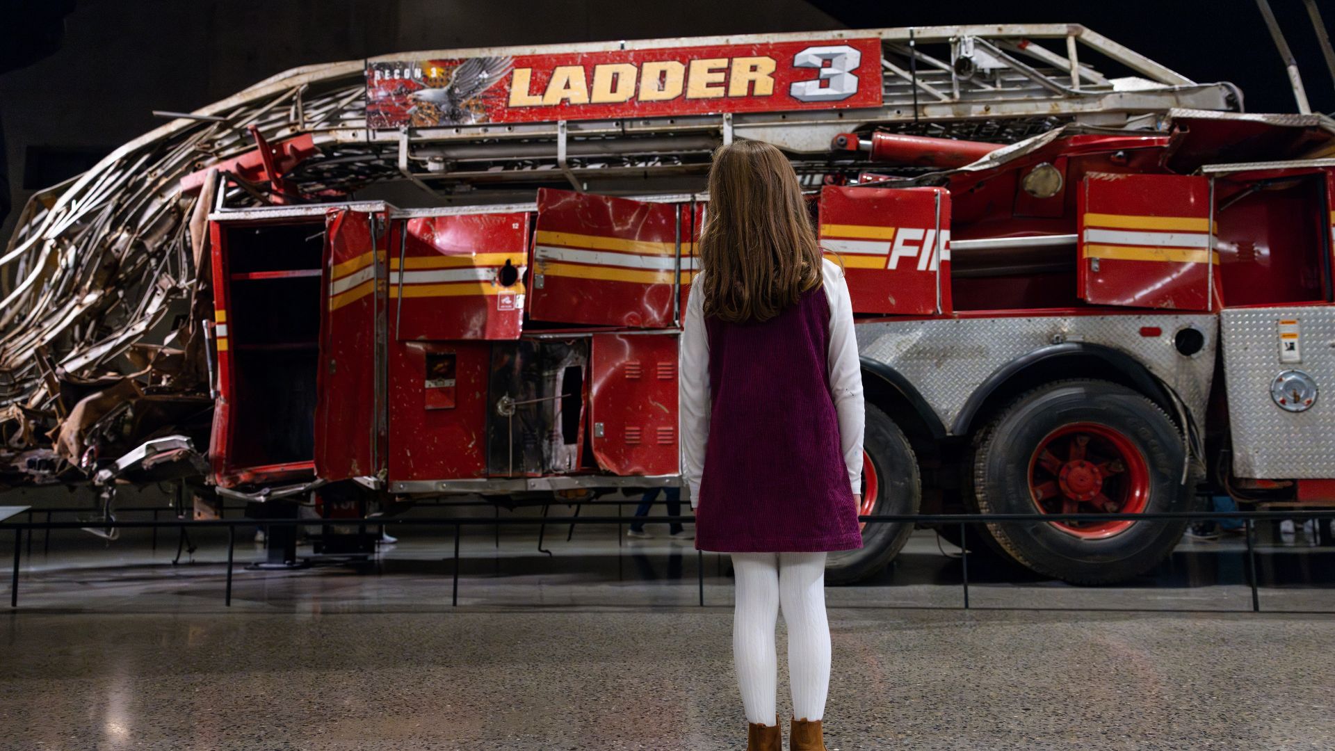 Back view of a child in a purple dress and white tights looking at a mangled fire truck 