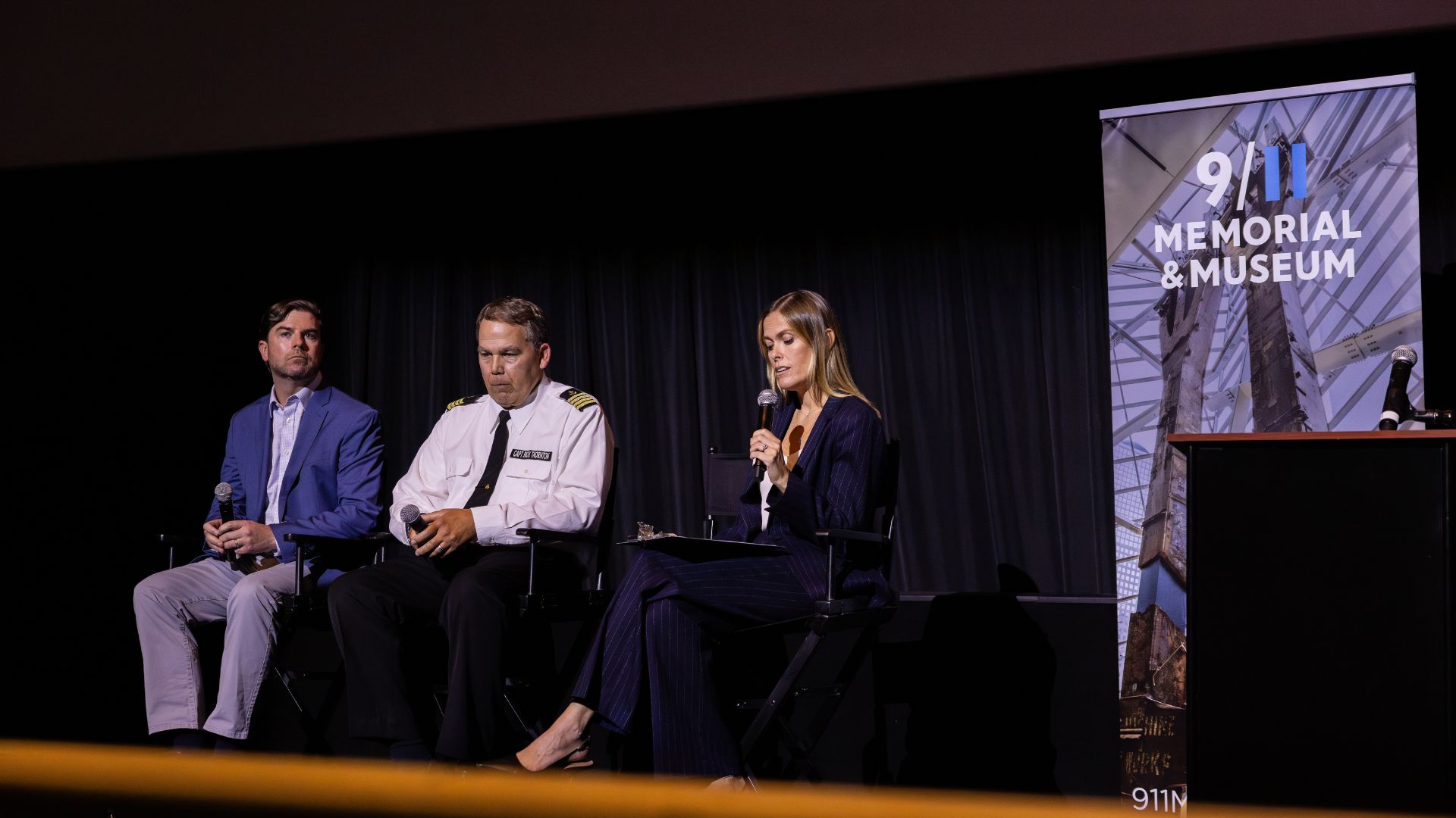 Three people seated, in conversation, on stage, with a dark backdrop behind them and a vertical banner that reads, "9/11 Memorial & Museum." Under the text is a photograph of the steel tridents.