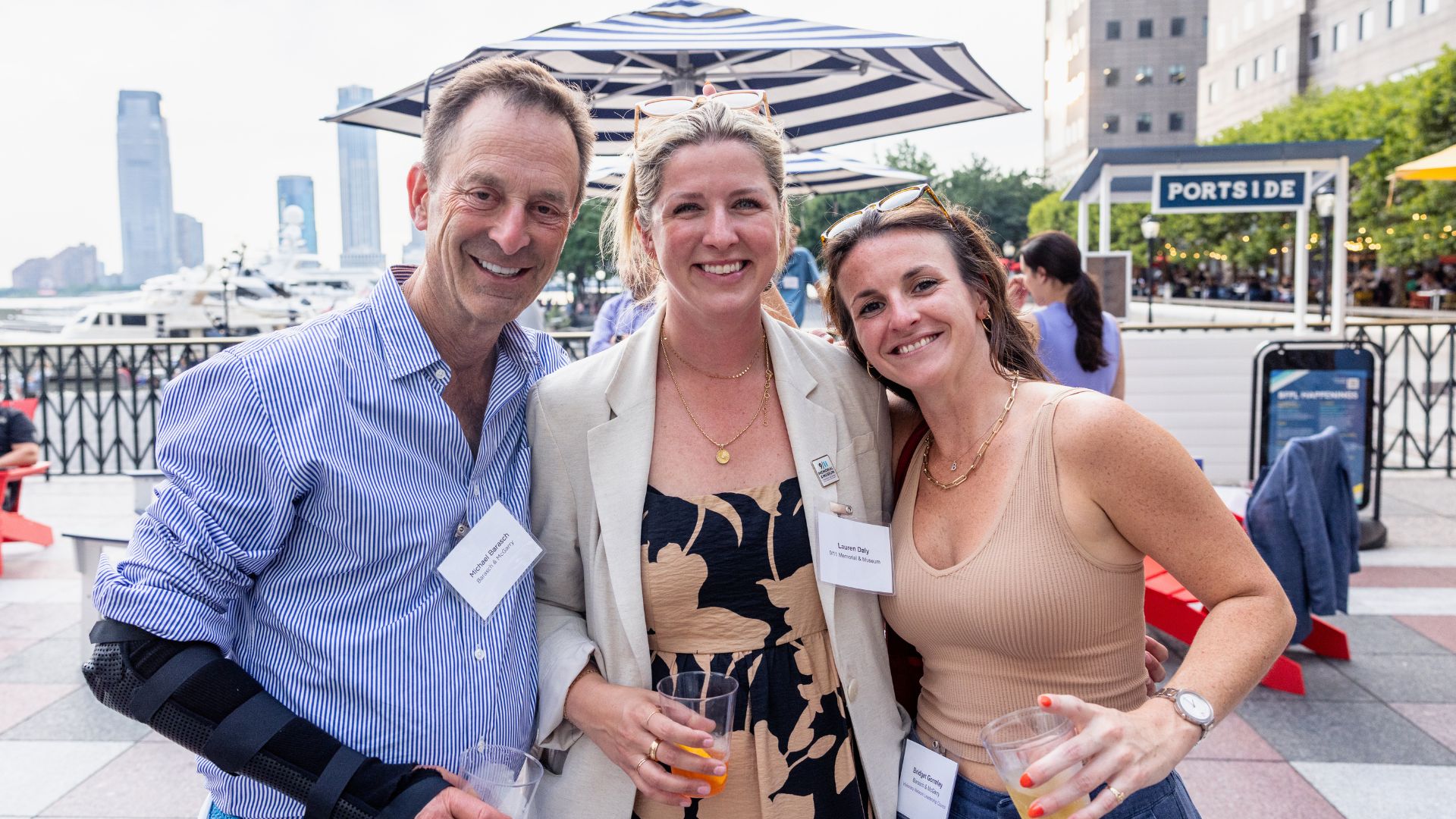 Three people smile as they pose with the Hudson riverfront behind them, and the top of a blue and white umbrella visible behind them.