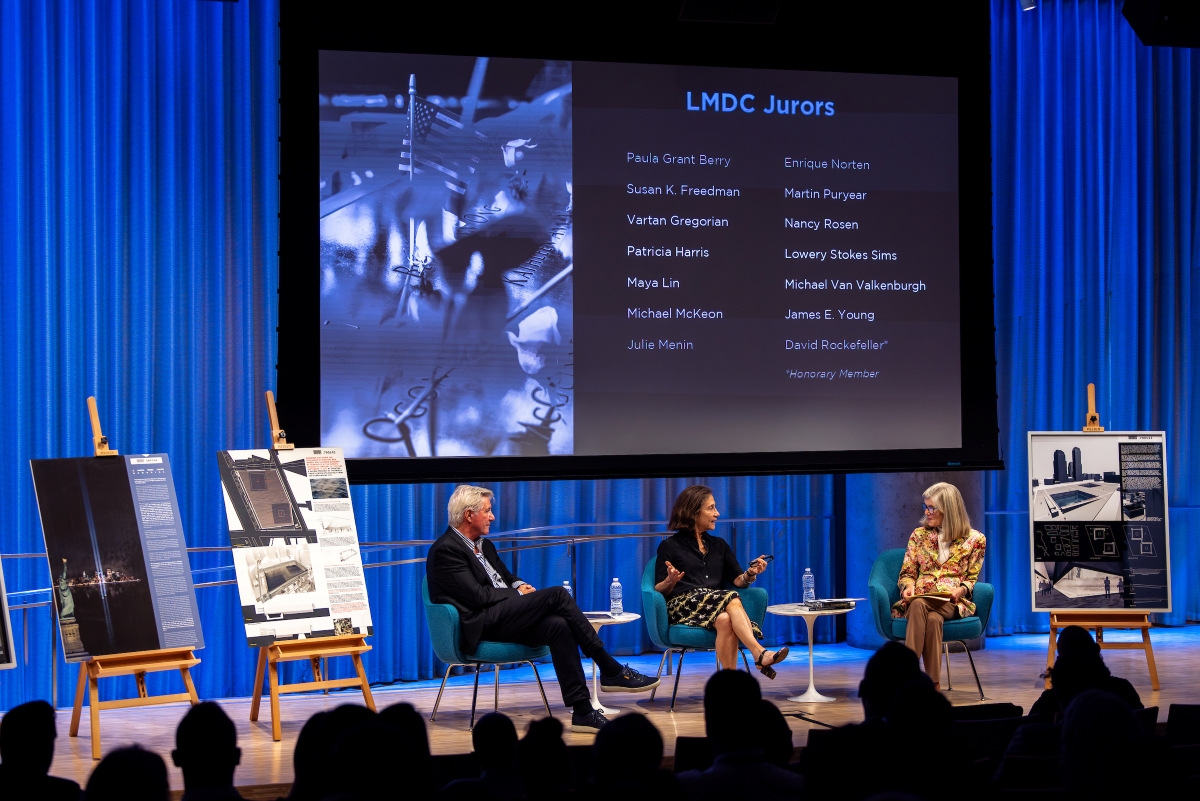 The full stage is visible in a wide shot with easel boards to the left and right of the panelists and a large screen with a digital image on the right and text on the left.