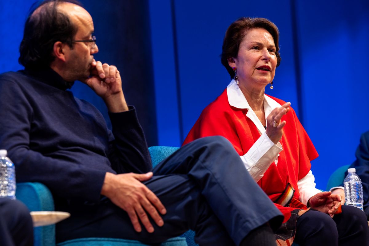 a woman in a red sweater speaks while a man beside her listens intently