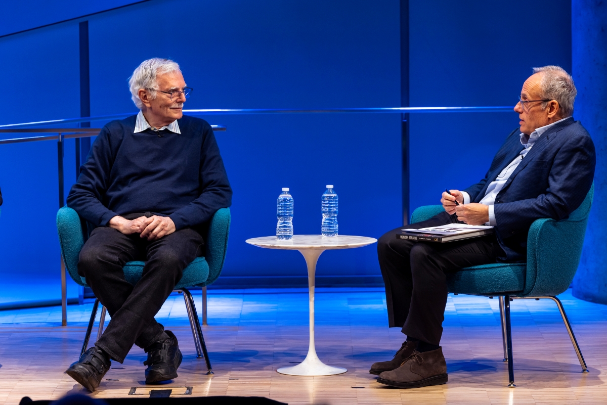 Two men sit on each side of a small white table, talking on a stage