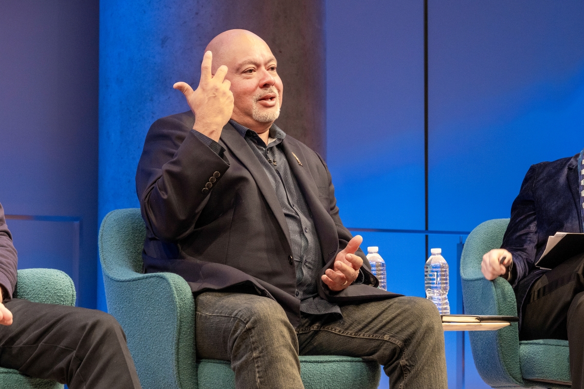 A man speaks and gestures with one had to his temple.