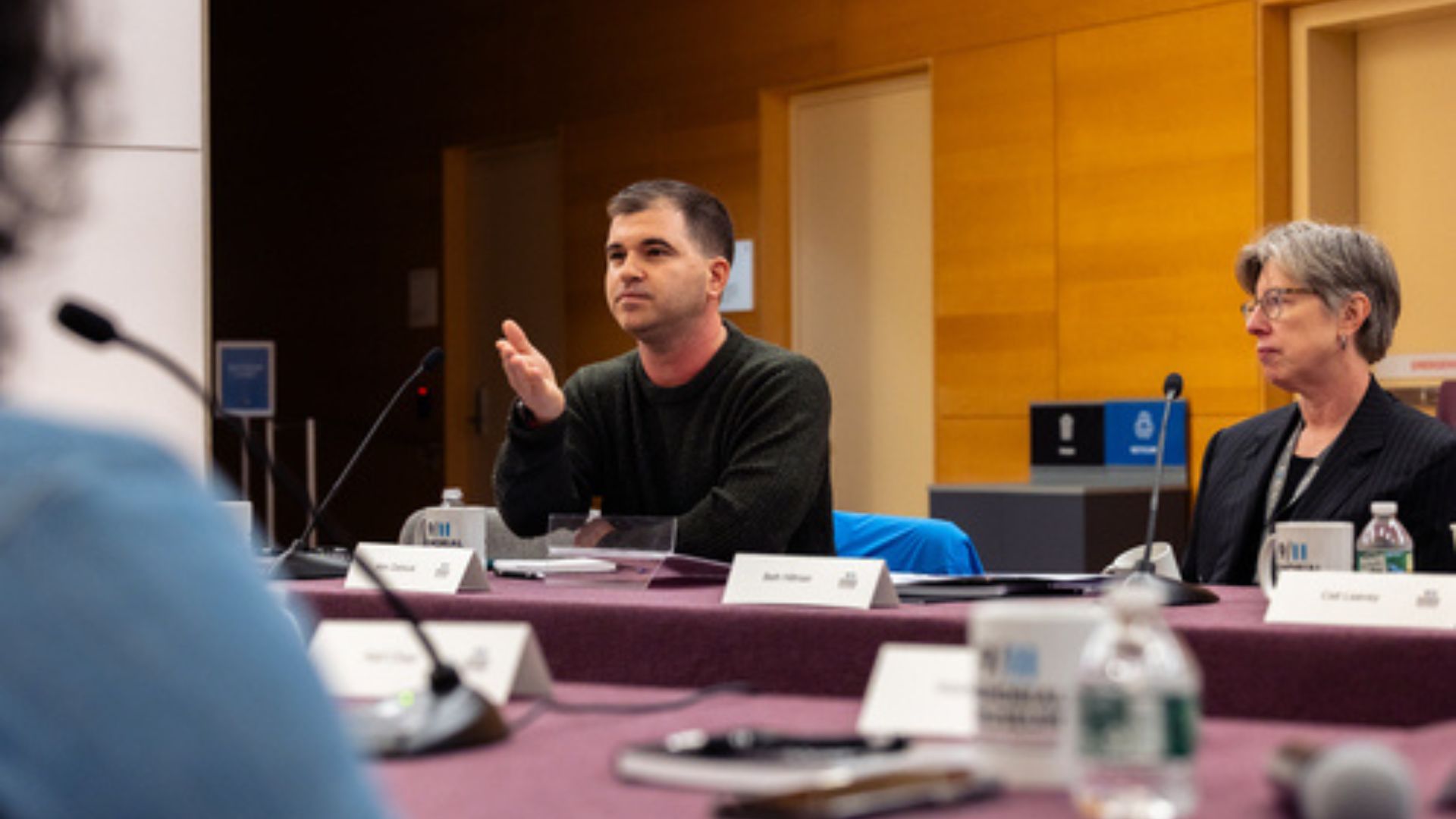 A speaker at a panel discussion is seated at a table while a woman to his left looks on.