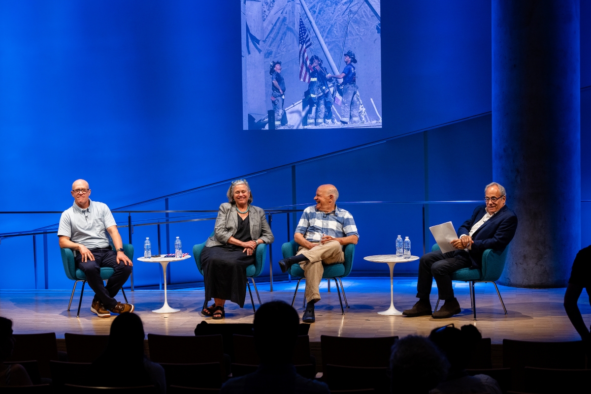four people sit on stage in front of a blue background with a large photo on the screen -- the flag from Ground Zero