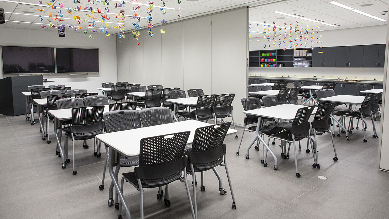 Black chairs and white tables fill an empty Education Center. There are two classrooms separated by a room divider. Colorful origami birds hang over the tables. Presentation screens and a lectern are at the front of the room. 