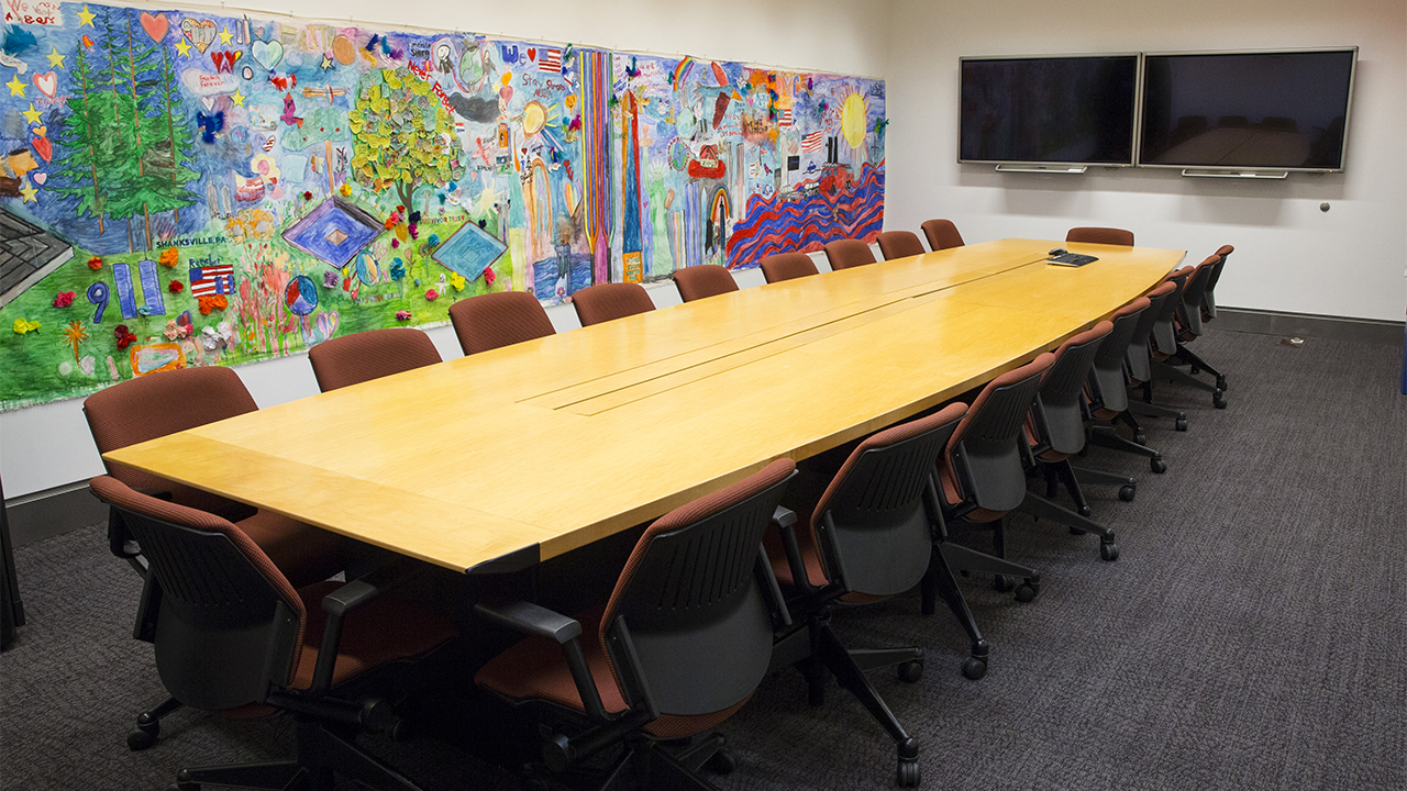 Red-cushioned rolling chairs surround a long, wooden conference table. A colorful mural of children’s artwork is on the wall. 