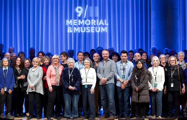 A crowd of about two dozen volunteers smile onstage at an event in the Auditorium. The logo of the 9/11 Memorial & Museum is projected on a blue stage curtain behind them. 
