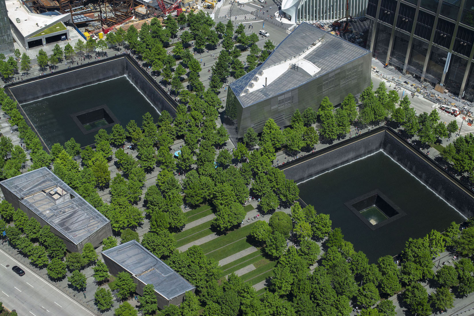 Memorial Plaza is seen from above on a sunny day. Dozens of oak trees fill the Plaza with a vivid green. These trees surround the two reflecting pools where the North and South towers once stood. Between the large, square pools is the Museum’s glass and steel pavilion. 