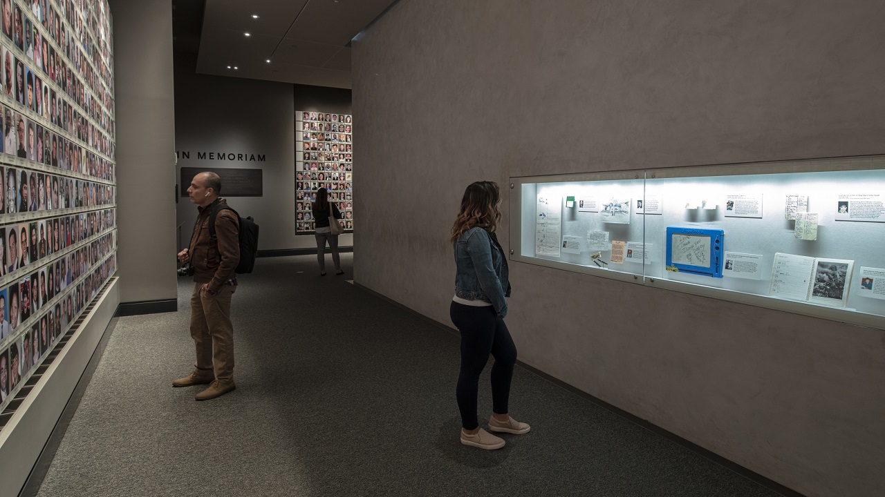 In this interior look at the In Memoriam exhibition, a visitor standing in the foreground is looking at personal items that belonged to victims of the attacks that are on view in a display case.  Two other visitors linger over some of the 2,983 portrait photographs of victims are on walls to the left and right of the chamber. 