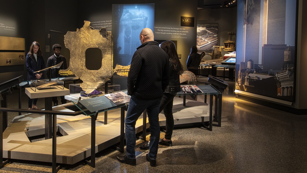 Artifacts from September 11, including a fire truck and pieces of an airplane, are on display at the Historical Exhibition. A lit image of the burning Twin Towers stands out at the center of the room. Other display areas and interactive screens are in the background. 