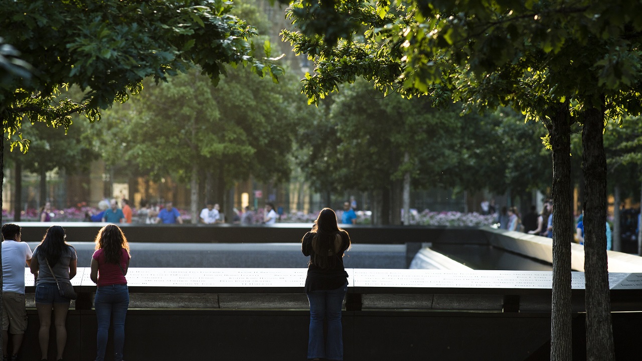 The back view of visitors looking at one of the Memorial pools, surrounded by green trees 